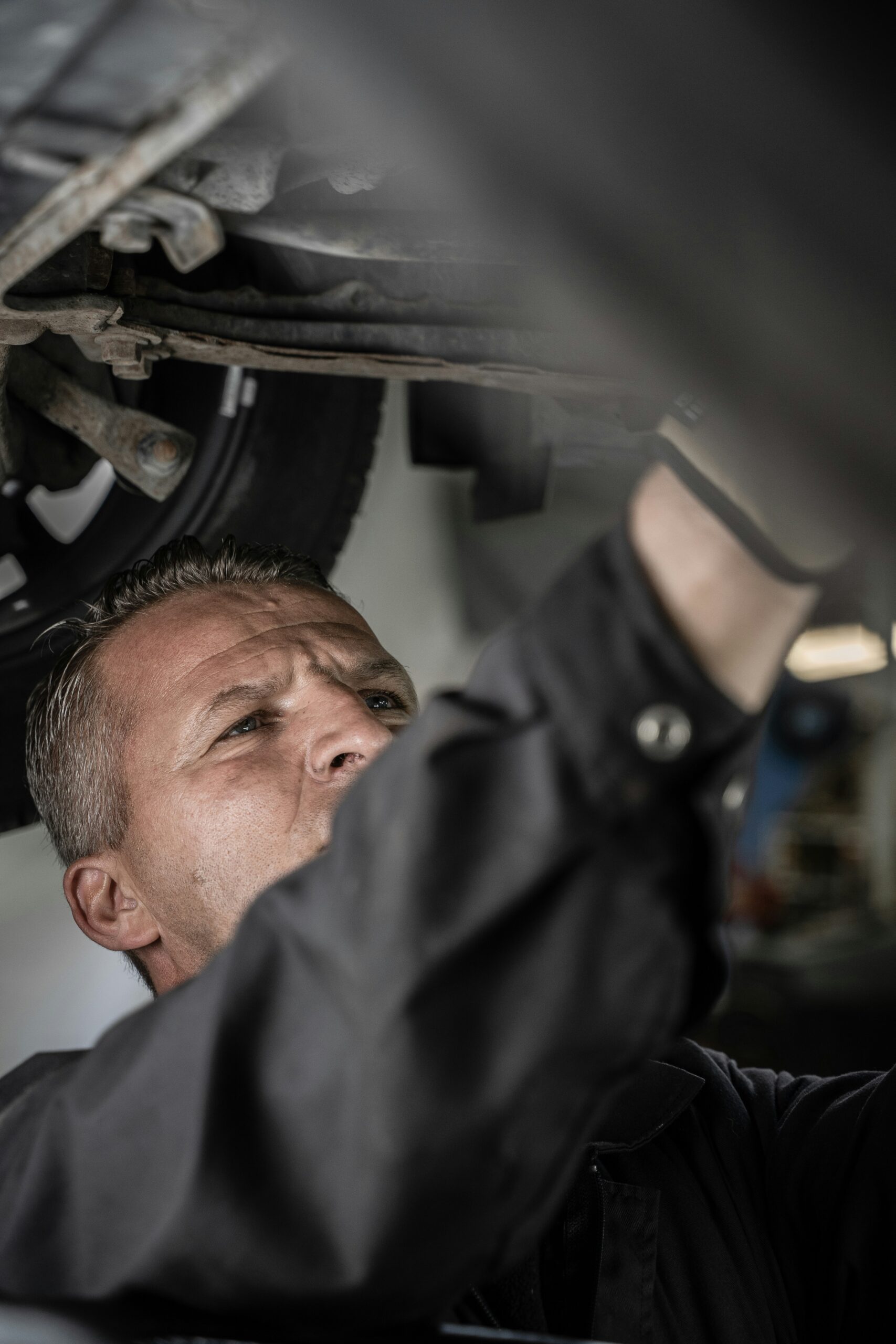 A mechanic wearing a dark work uniform inspects or repairs the underside of a vehicle, focusing intently on his task. The photo is taken from a low angle, showing the cars undercarriage and his concentrated expression.