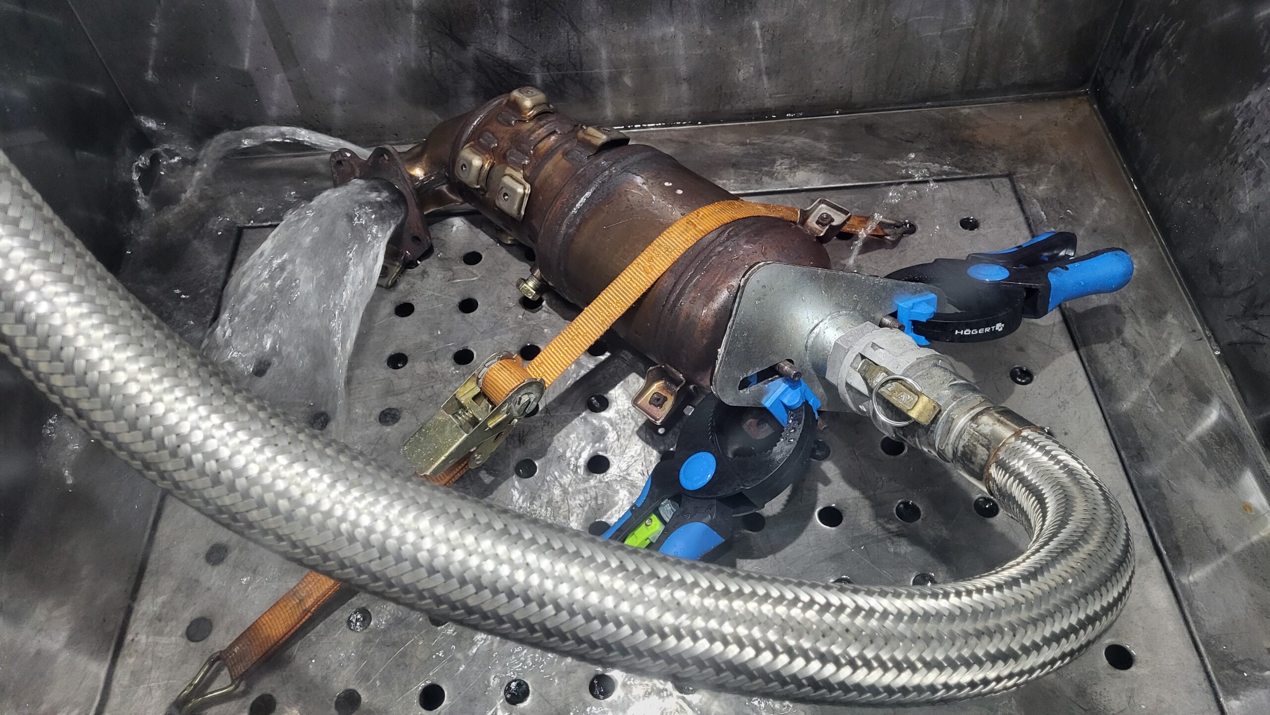 A large cylindrical metal industrial component with various ports, pipes, and attached control units sits on a worn concrete floor next to a wooden pallet.