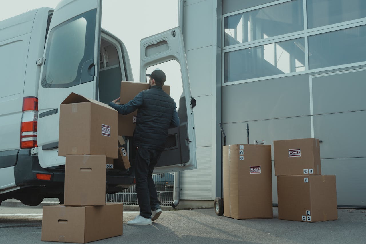 A person wearing a jacket and cap is unloading large cardboard boxes from a white delivery van parked outside a building. Several boxes are stacked on the ground nearby.